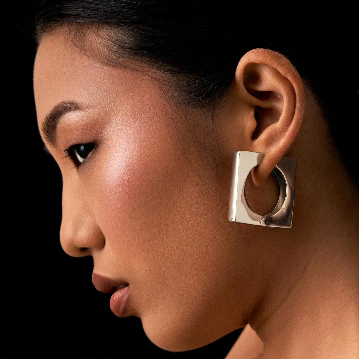 Fotografia editorial de moda com perfil em close-up de uma modelo com cabelo escuro preso, usando a Argola Quadrada Bold com design geométrico e banho de ródio premium da Ahal Acessórios. A imagem com iluminação contrastante destaca perfeitamente a escala real de 3,5cm, o caimento impecável e o luxo contemporâneo da peça na orelha.