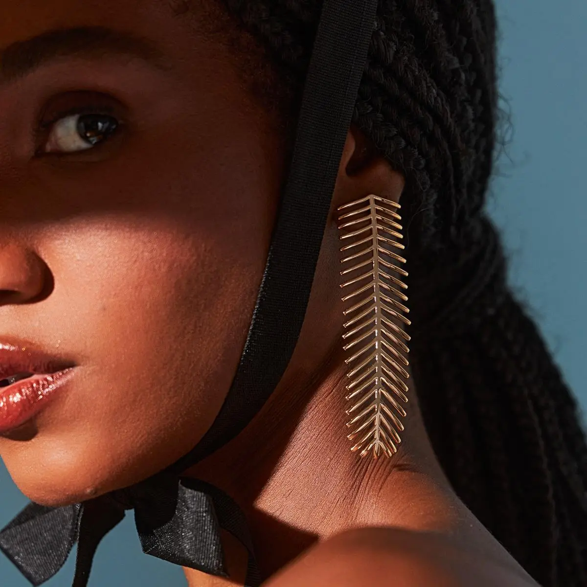 Modelo de pele preta de perfil com cabelo em rabo de cavalo com tranças usando o Brinco Longo Folha Espetada Liso Luxuoso Banho Ouro 18k em estúdio com fundo azul céu.