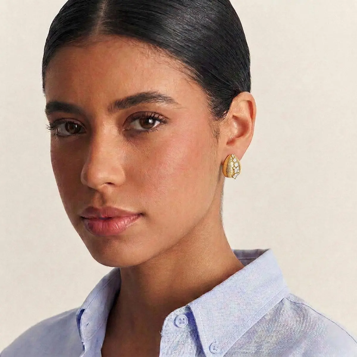 Modelo de pele parda e cabelo preto preso em coque usando o Brinco Gota Texturizada Delicado com Cristais e Zircônias Banho Ouro 18k em fundo branco.