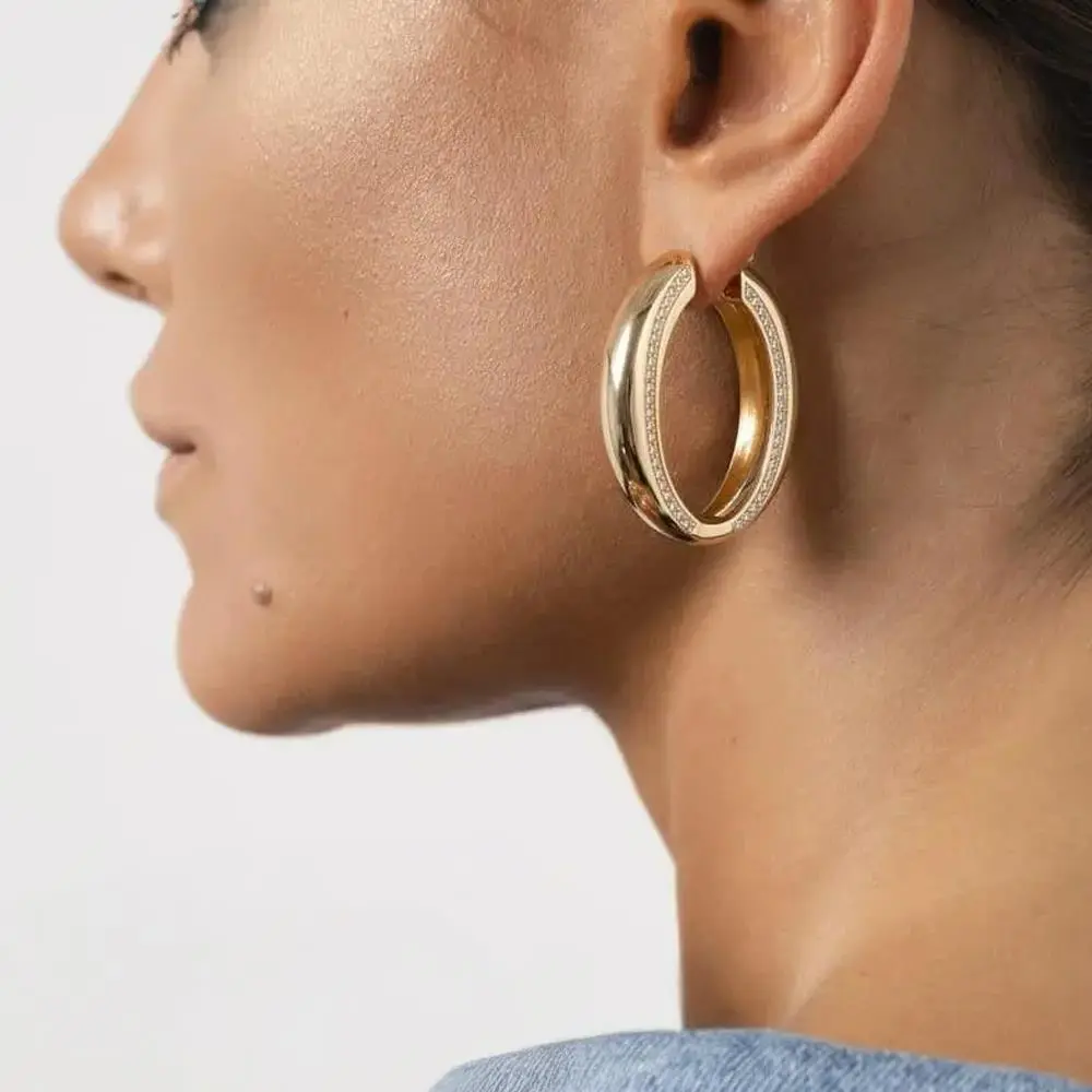 Foto de perfil em fundo claro destacando brinco argola oval grossa com zircônias na orelha da modelo.