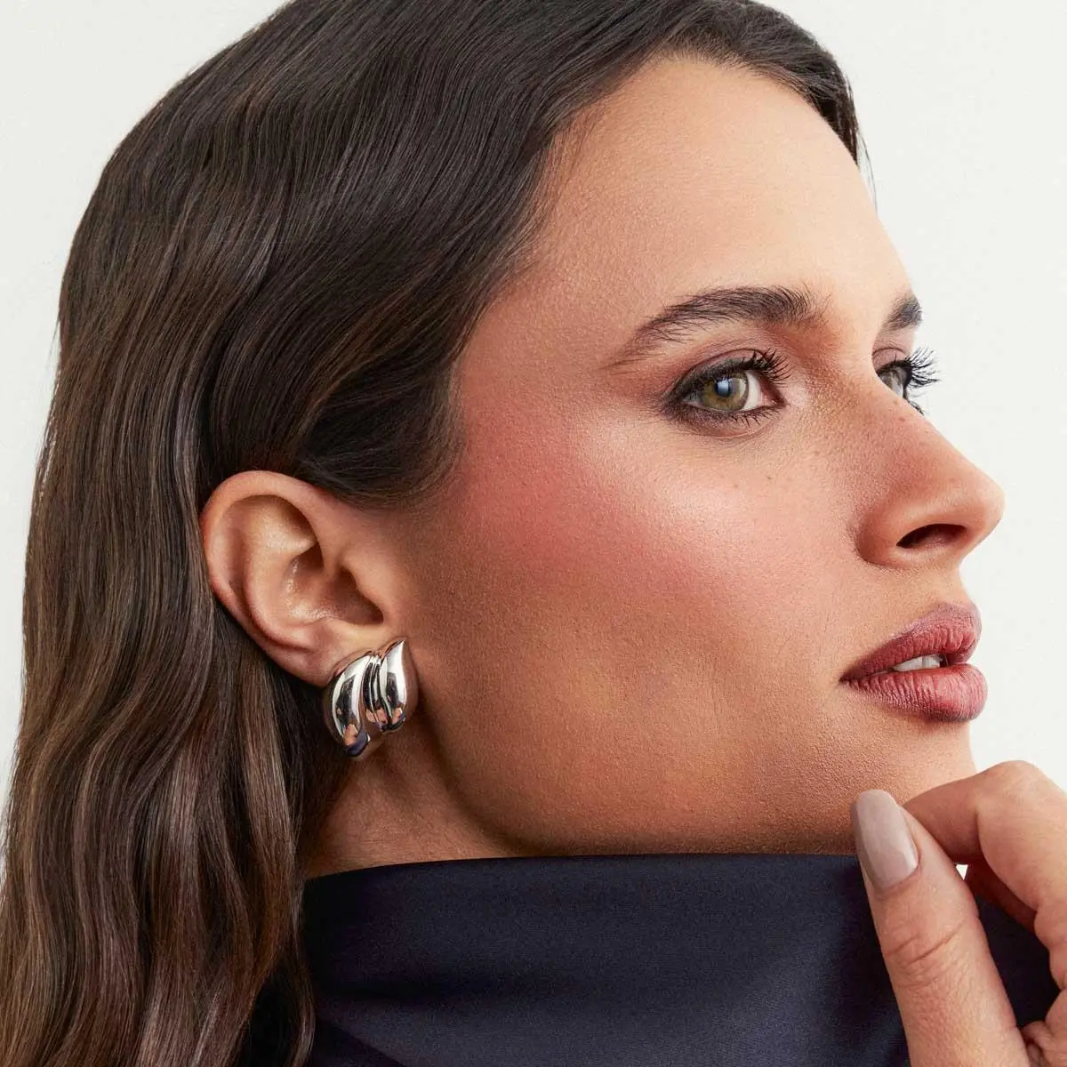 Fotografia editorial em estúdio com foco de perfil em close-up de uma modelo de pele clara e cabelos castanhos claros presos. Ela usa o Brinco Concha Abaulado Liso com design orgânico e banho de ródio premium da Ahal Acessórios. O enquadramento destaca perfeitamente a escala real, o caimento fluido e o brilho espelhado da semijoia de luxo aplicada à orelha, contrastando com sua blusa escura de gola alta.
